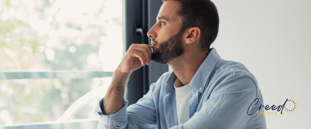 Thoughtful man in a light blue shirt looking out a bright window with his hand resting on his chin